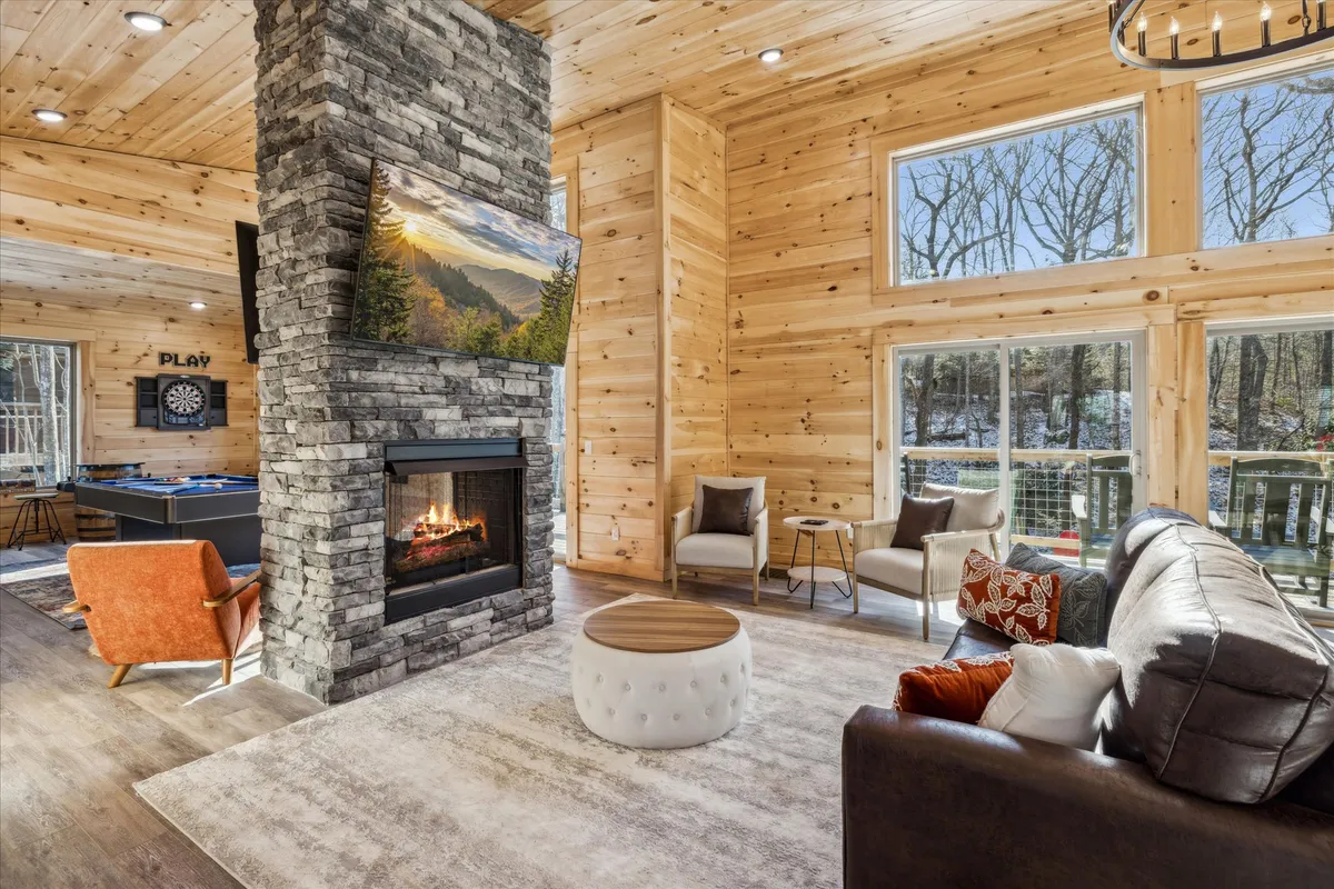 A spacious living area with a grand stone fireplace, generous seating, and expansive windows letting in plentiful natural light. The setting encourages relaxation, connection, and socializing amidst a warm, welcoming atmosphere.