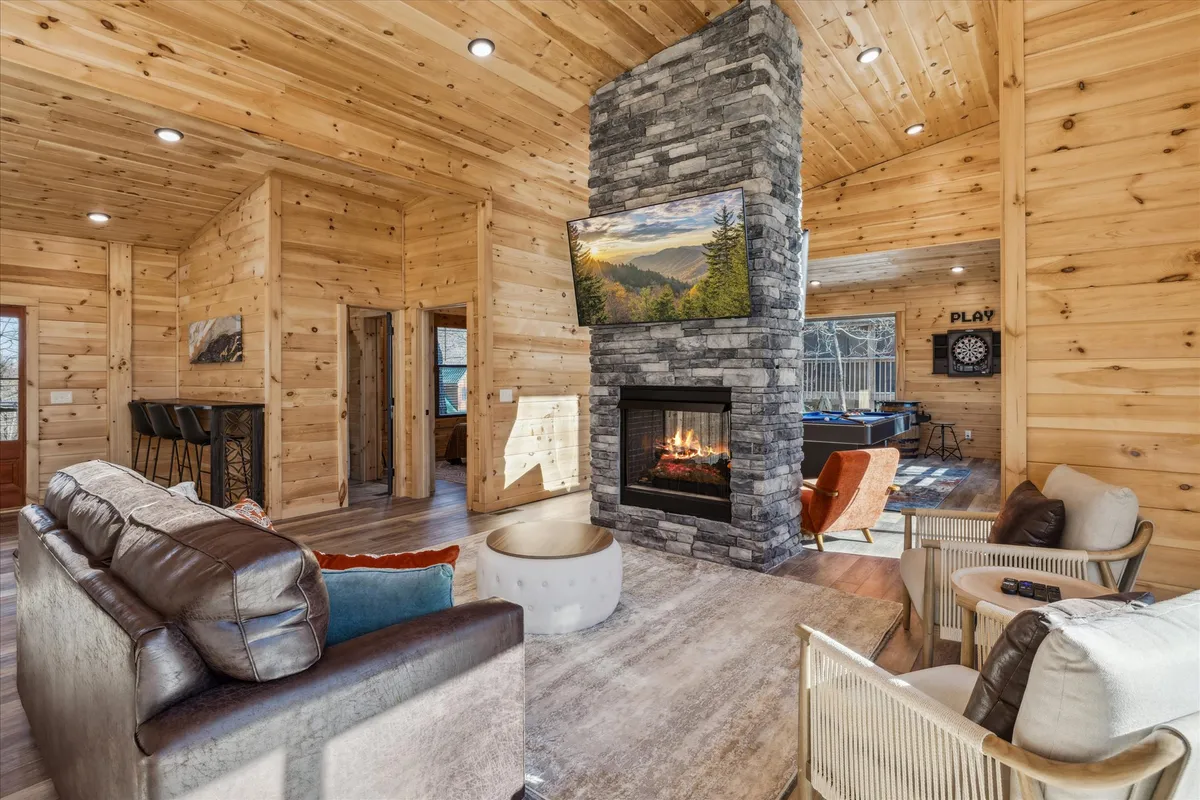 The image shows a cozy living area featuring a striking stone fireplace, a TV, and large windows that frame mountain views. The wooden walls and designer touches create a warm, inviting atmosphere that feels luxurious yet intimate.