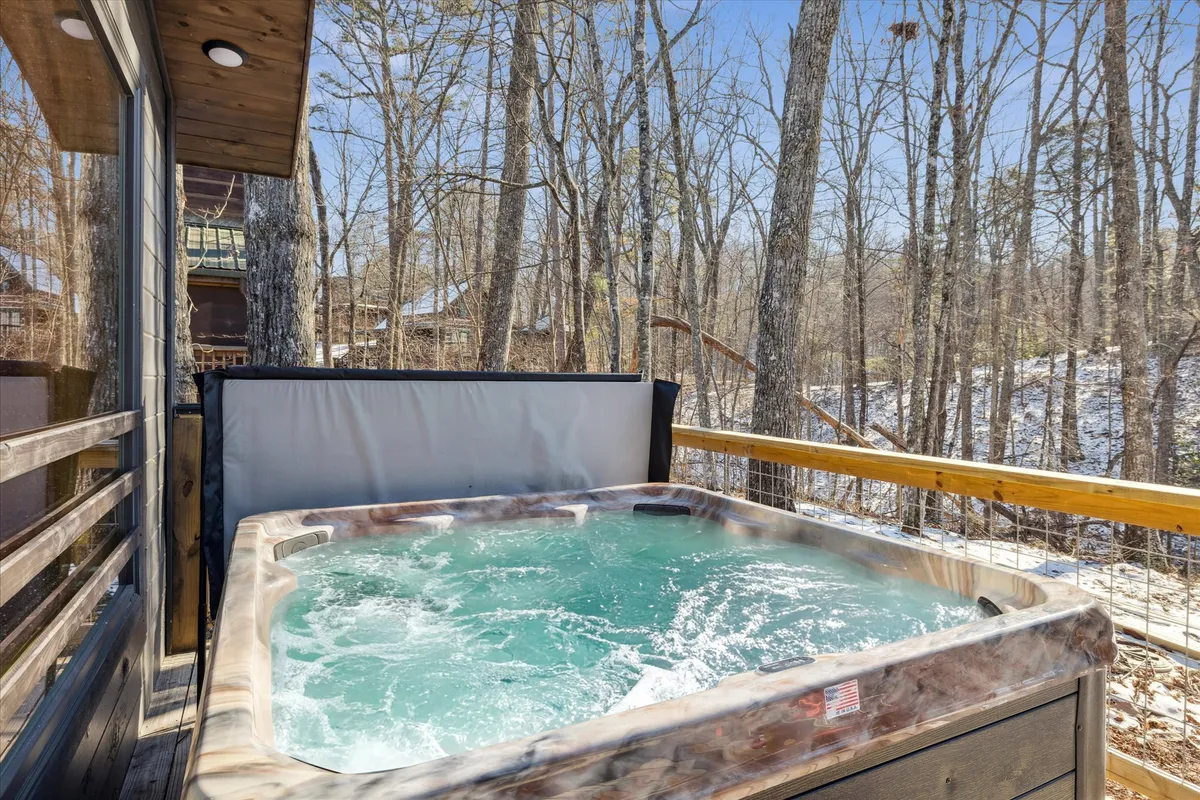 An outdoor hot tub sits on a private deck surrounded by trees, with a sturdy railing and tranquil views of nature. The space is designed for relaxation, offering guests a soothing environment to unwind under the open sky—ideal for stargazing or soaking after a day of games.