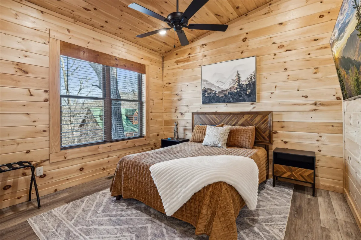 This scene focuses on a bed with a decorative throw, artwork above the headboard, and large windows looking out onto the surrounding landscape. The setting is serene and intimate, with soft natural light and comfortable furnishings.