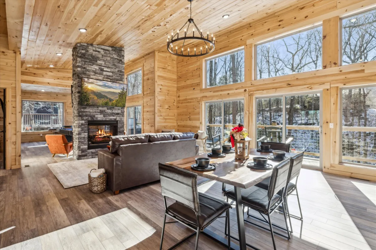A warm, inviting living and dining area with a stone fireplace, large windows framing the forest outside, a spacious wooden dining table, and a comfortable sofa. The natural light and open design create an atmosphere perfect for gatherings and relaxation.