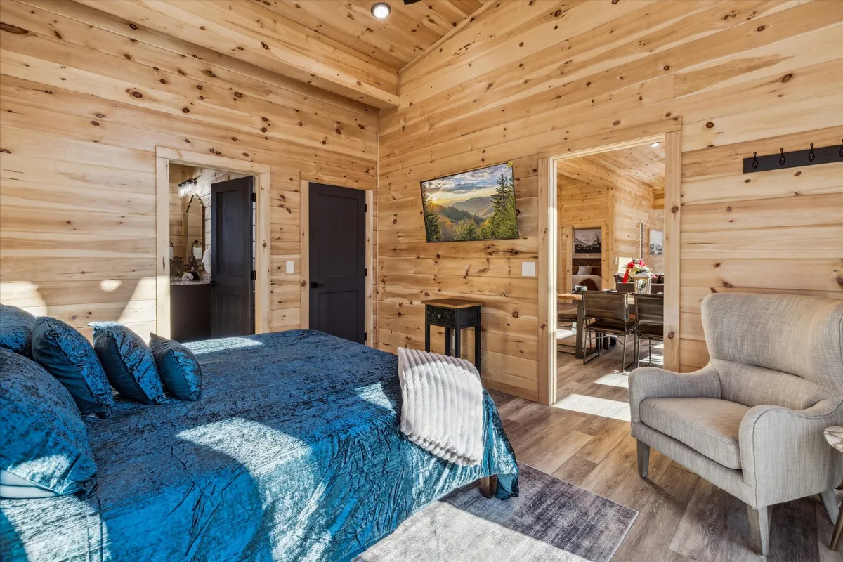 A serene bedroom featuring plush teal bedding, warm wooden walls, elegant artwork, and a comfortable armchair by the window. The window frames views of the surrounding natural landscape, enhancing the sense of peaceful seclusion.