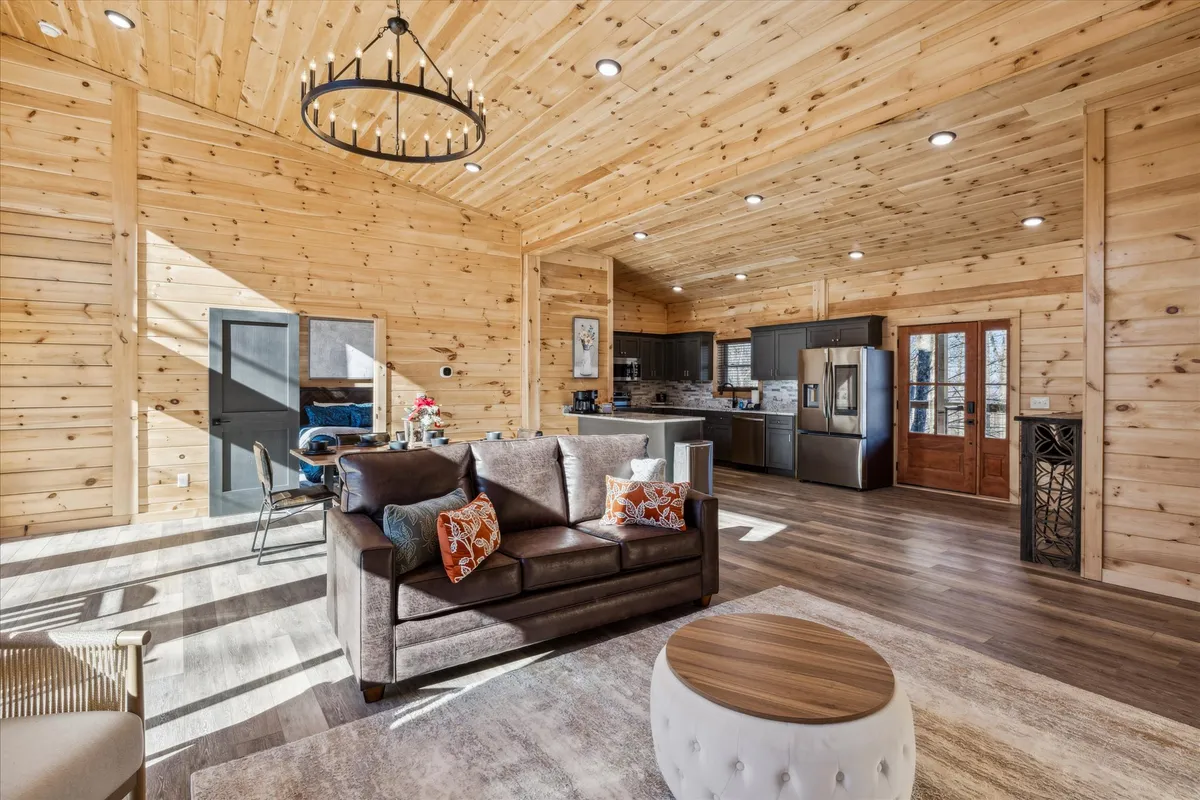 A wide view of the open living, dining, and kitchen area, featuring a comfortable sofa with colorful cushions, a wooden dining table with chairs, and visible kitchen space in the background. Soft natural light pours in from the windows, emphasizing the welcoming layout.