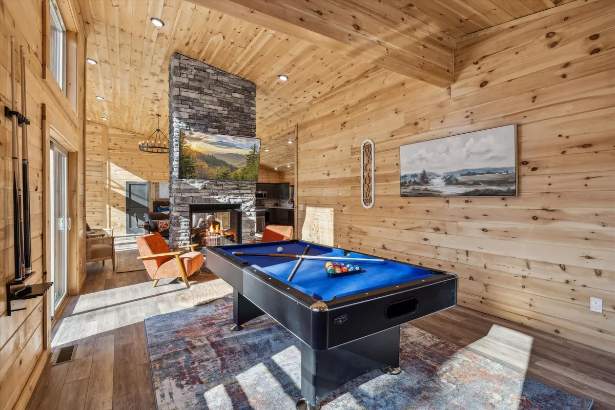 The image shows a spacious game room with a pool table as its centerpiece, flanked by a cozy stone fireplace and comfortable seating. Large windows allow in plenty of natural light and offer scenic views of the surroundings, enhancing the inviting, recreational ambiance. The room is styled with warm wooden accents that create a welcoming and upscale atmosphere.