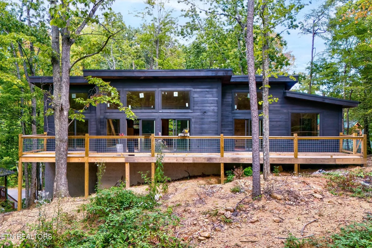 A modern cabin exterior surrounded by tall trees, with large windows and an expansive deck area. The natural setting suggests immediate access to the outdoors and potential for wildlife sightings or starting off on nature walks.