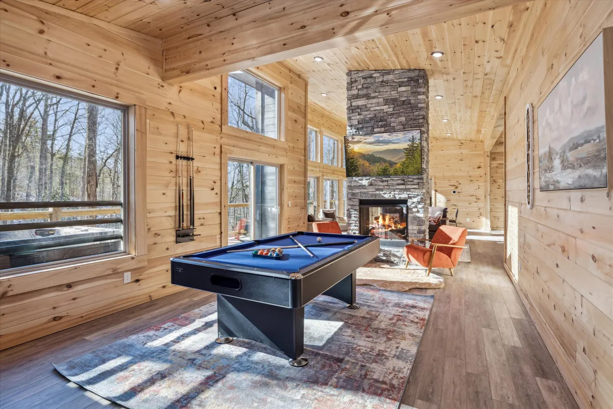 A vibrant game room featuring a pool table at the center, flanked by a cozy fireplace, large windows that frame scenic views, and comfortable seating. The space is bathed in natural light, inviting guests to gather, play, and socialize.