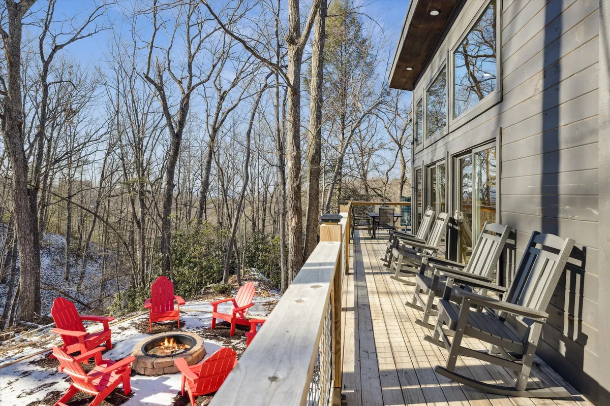 This exterior image features inviting red Adirondack chairs placed around a fire pit on a spacious wooden deck, with large windows of the cabin visible in the background and a glimpse of the surrounding natural environment.