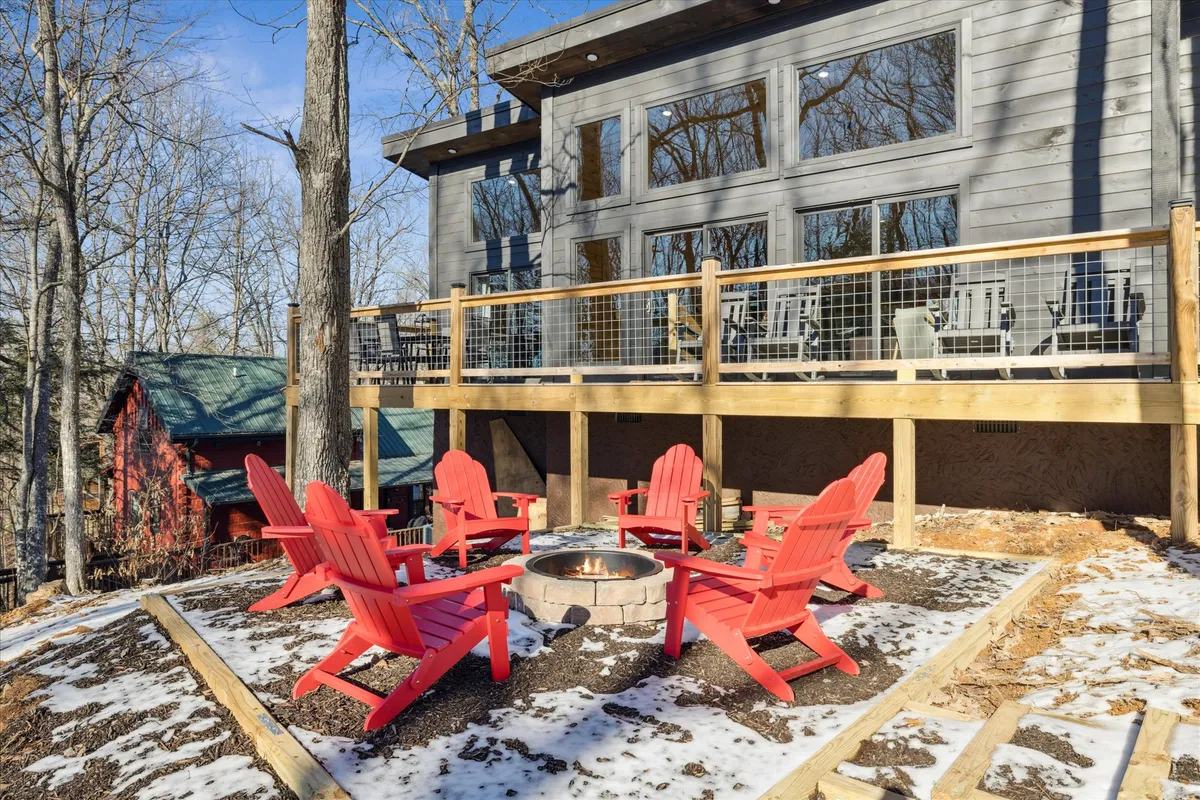This image features a cozy outdoor fire pit area surrounded by red Adirondack chairs, with large windows of the cabin and lush woodland in the background. The inviting setup signals a perfect spot for gathering, storytelling, and stargazing in a tranquil mountain atmosphere.